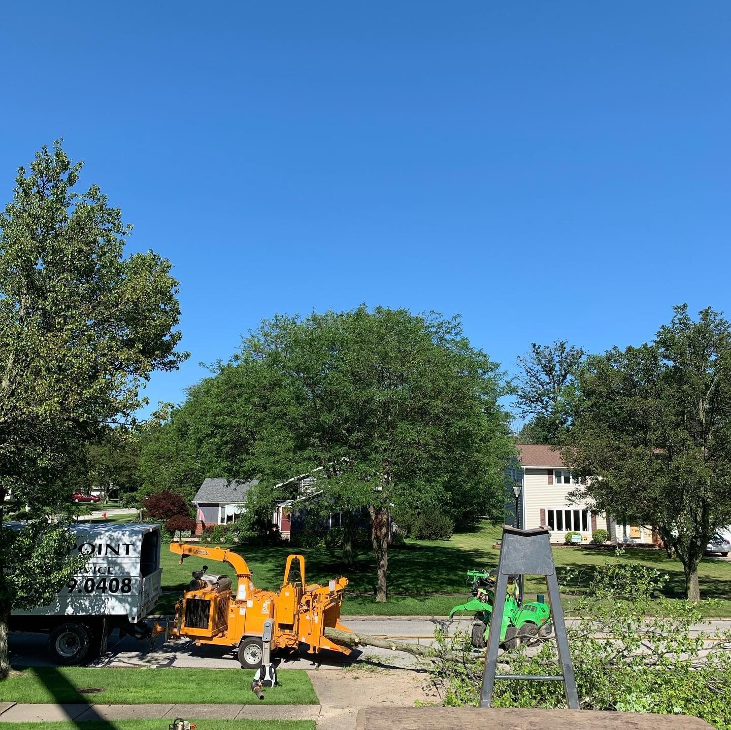 A white truck and bright yellow wood chipper parked on a suburban street in front of green trees and a two-story home.