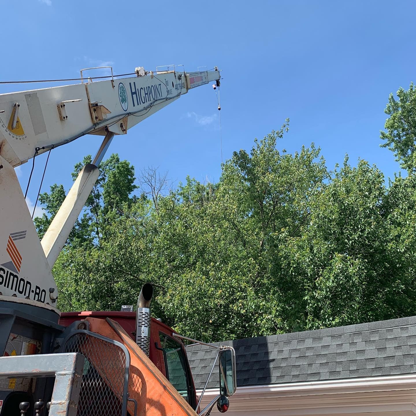 A crane arm extends against a bright blue sky above a residential rooftop and green trees.