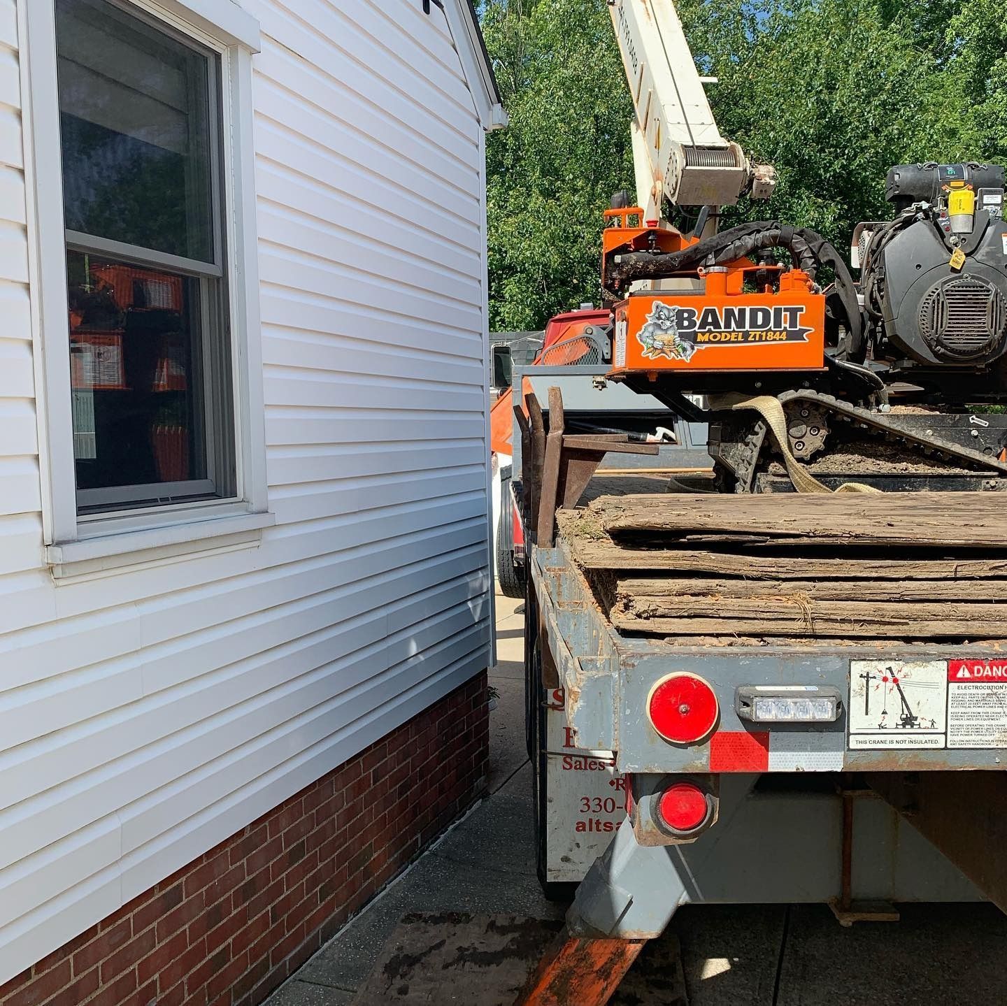 An orange Bandit brand track-mounted wood chipper sits on a trailer parked beside the brick and white-sided wall of a house.