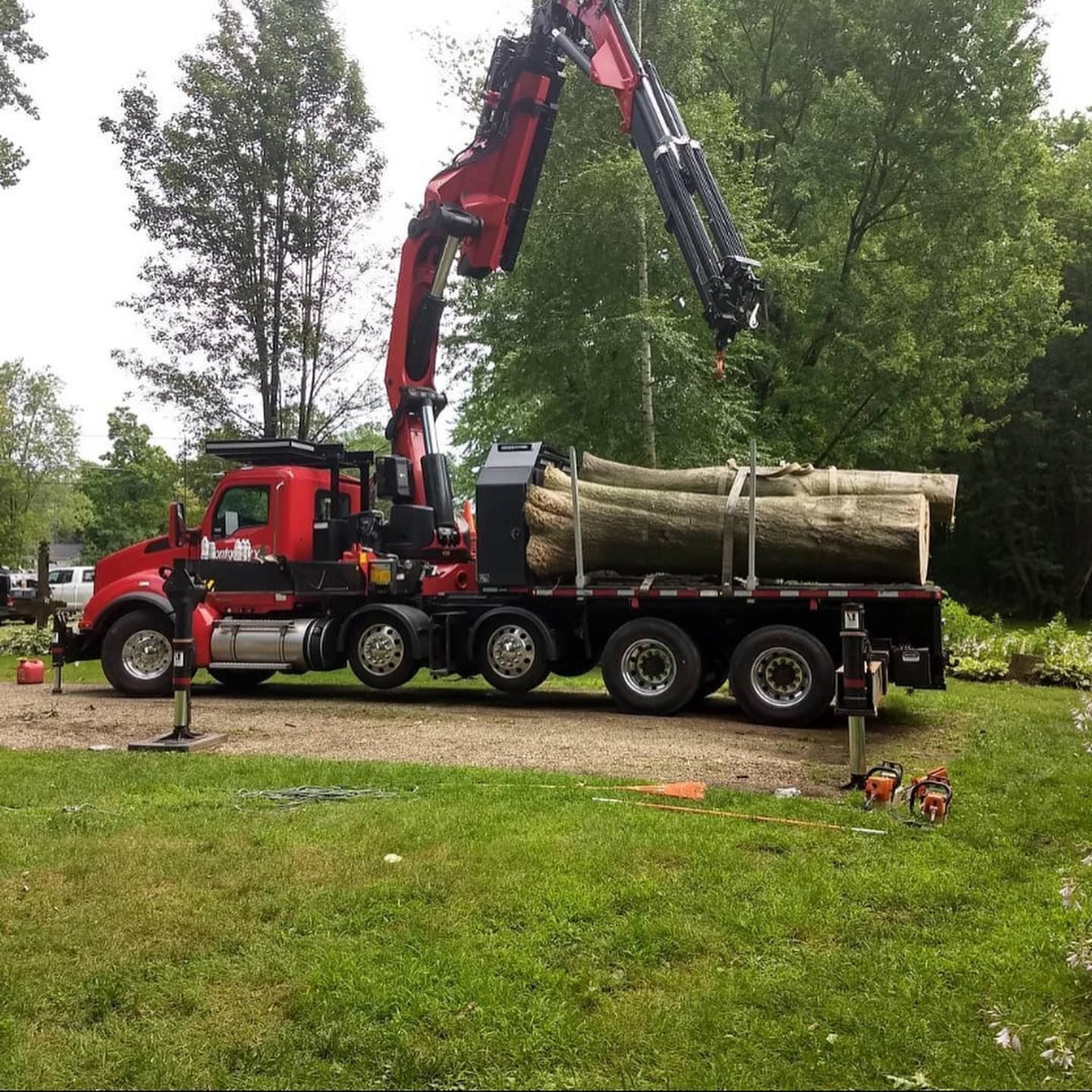 A red flatbed truck with a hydraulic crane and large logs parked on a grassy lot with trees in the background.