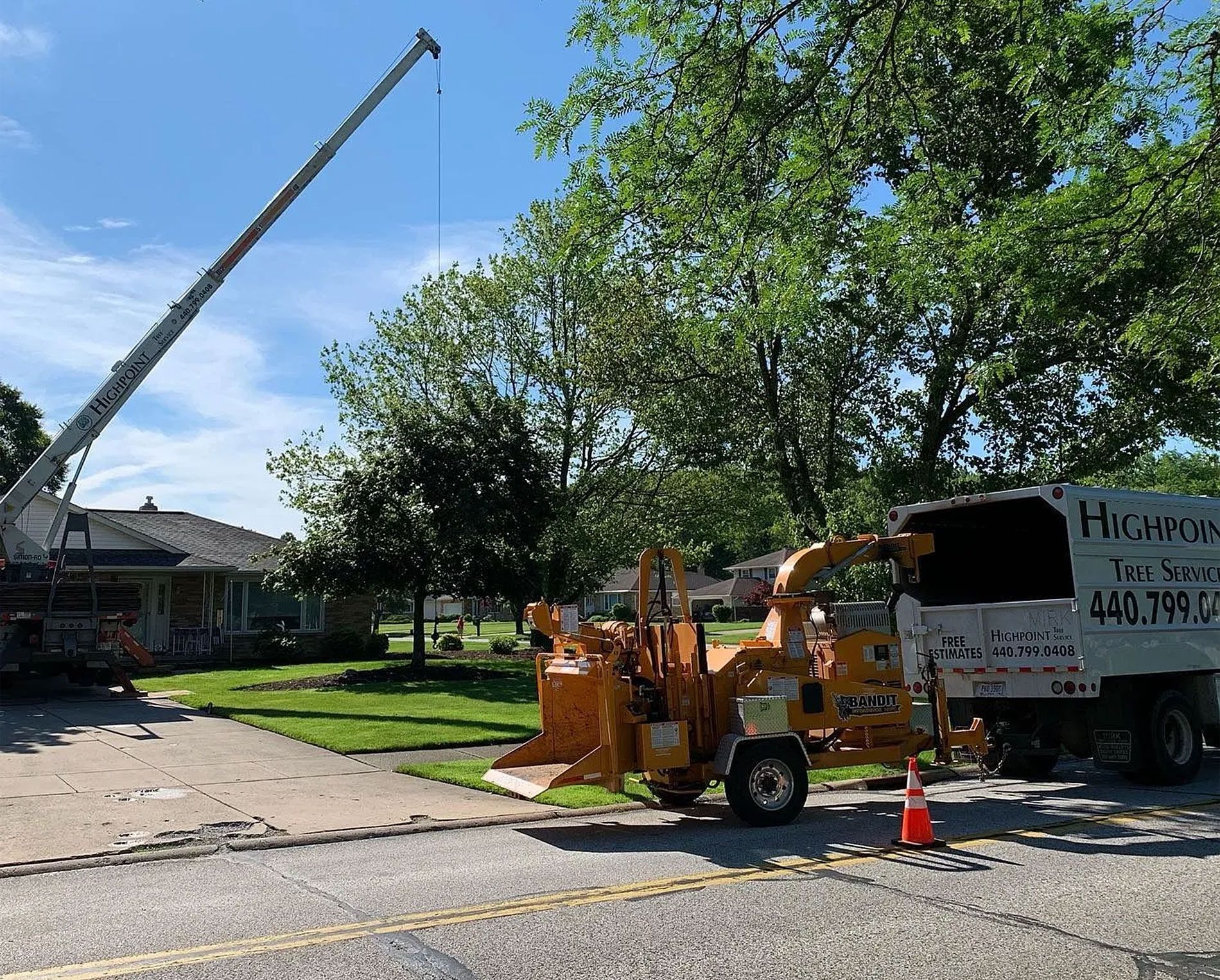 A crane and wood chipper parked on a suburban street in front of a house and mature trees under a bright blue sky.