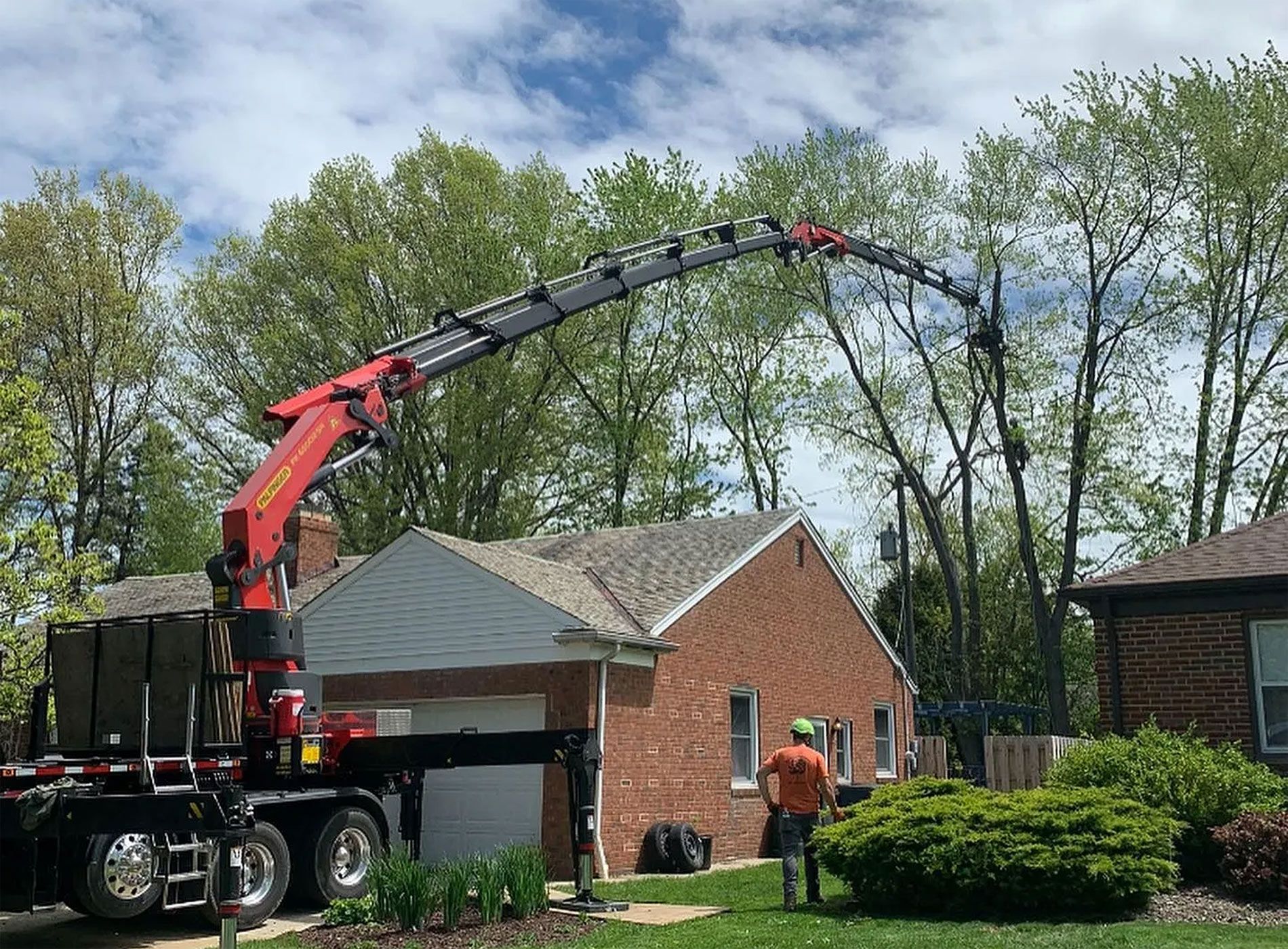 A red knuckle boom crane parked near a brick house extends its long, segmented arm toward tall trees for trimming.