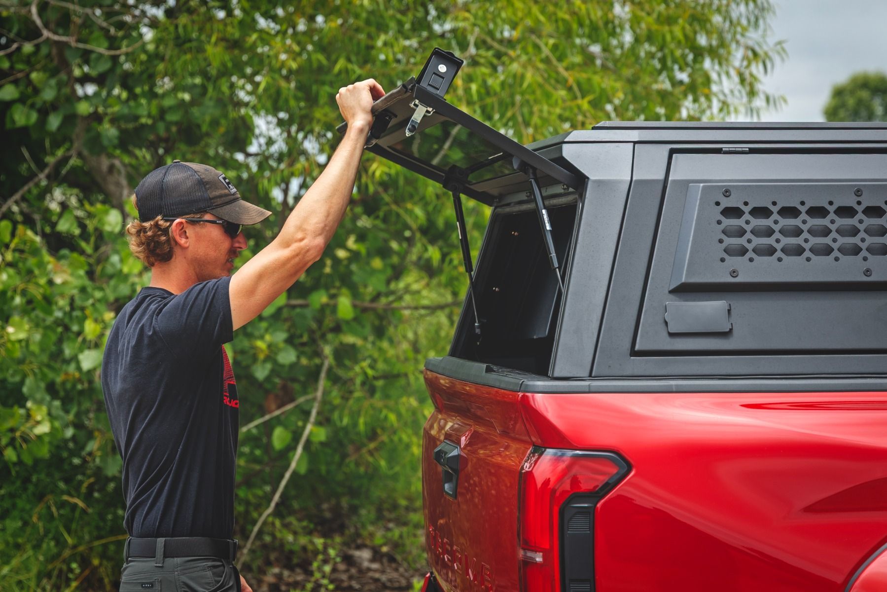 Man opening the rear canopy of a red pickup truck outdoors in a wooded area