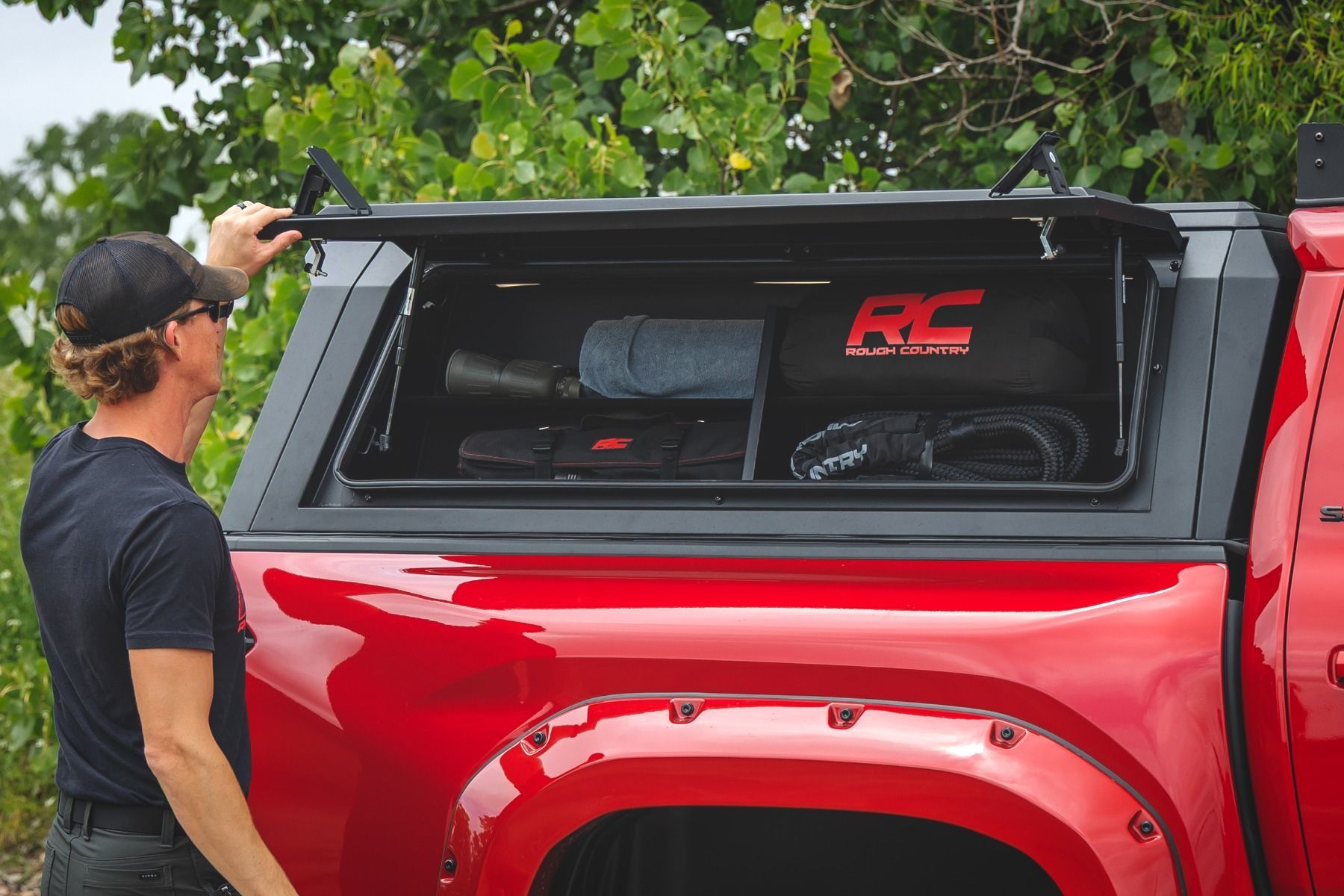 Person standing by a red pickup truck with a raised tonneau cover, inspecting the bed area.