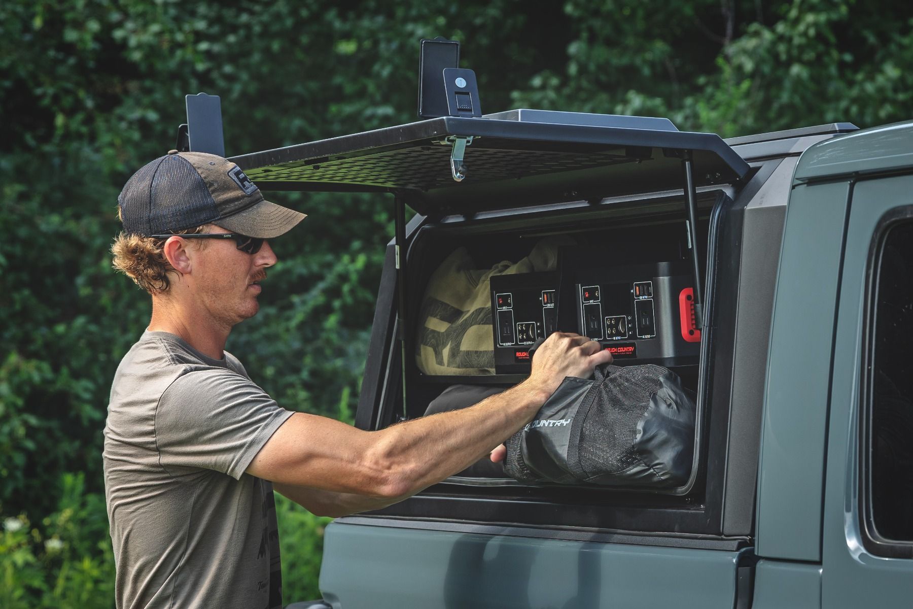 Man loading gear into a green pickup truck with an open canopy in a wooded area