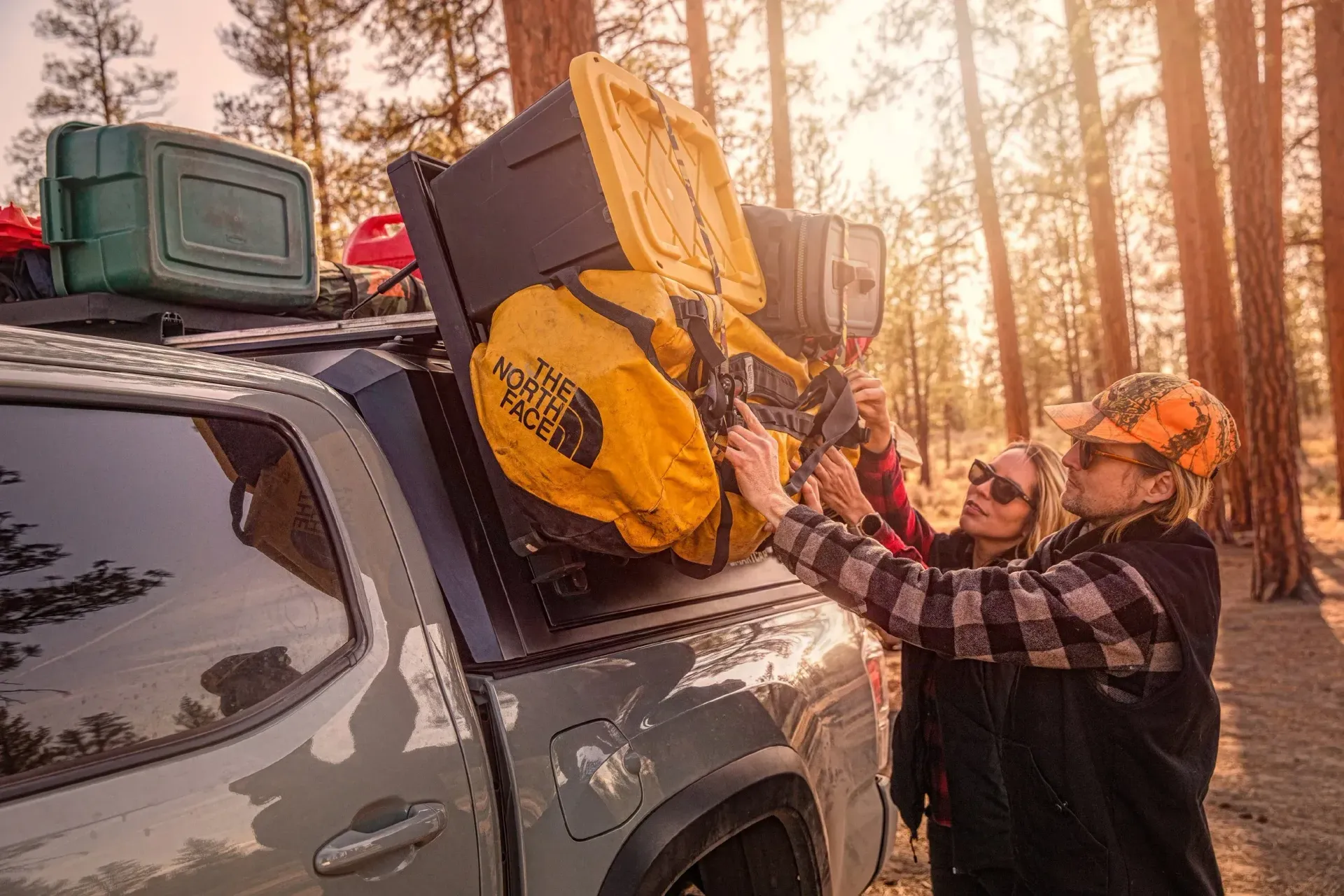 Three people loading gear onto a truck in a sunny forest setting. Bright yellow bags and containers on the rack.