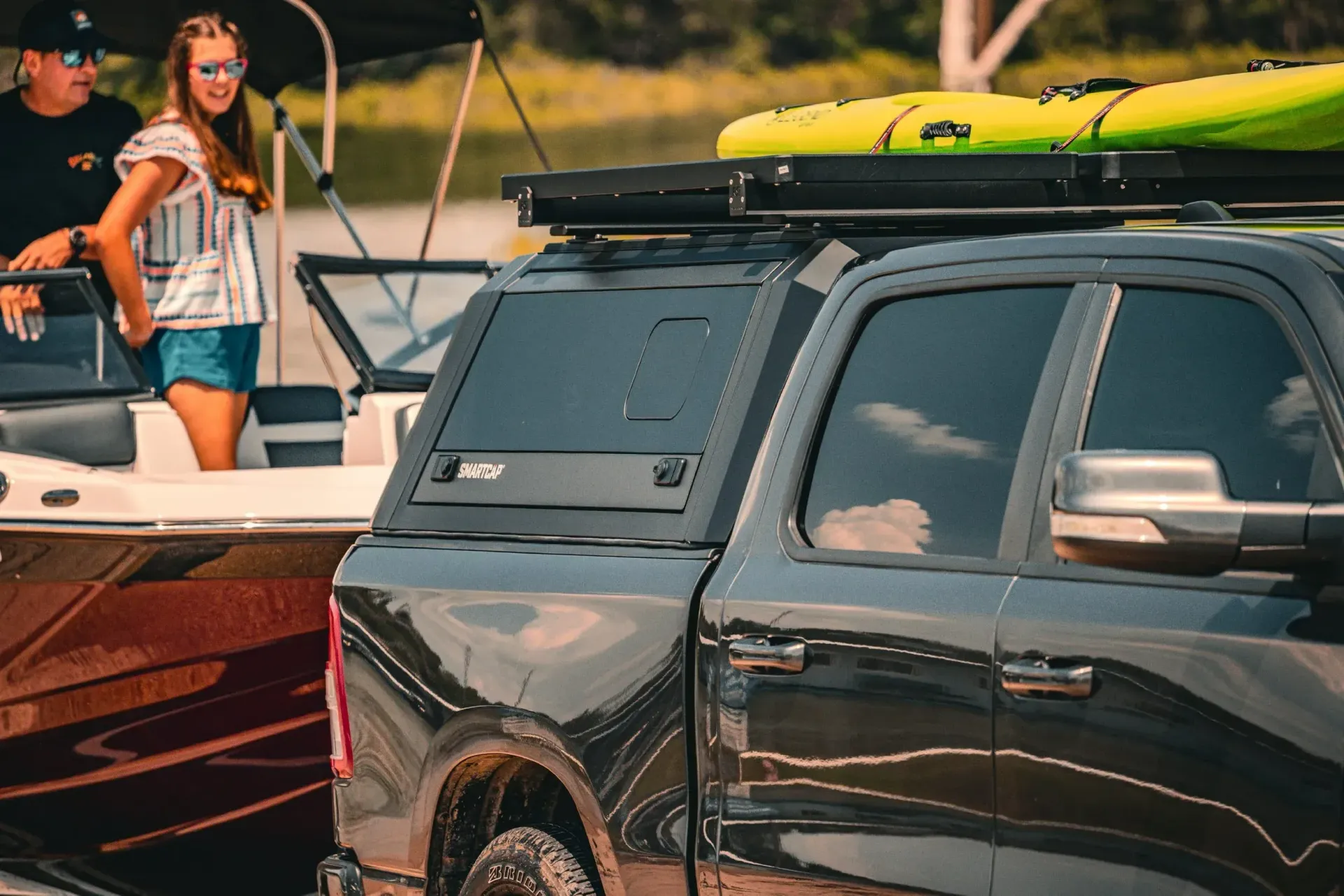 Black truck with camper shell, two kayaks on the roof, near a boat at a lake.