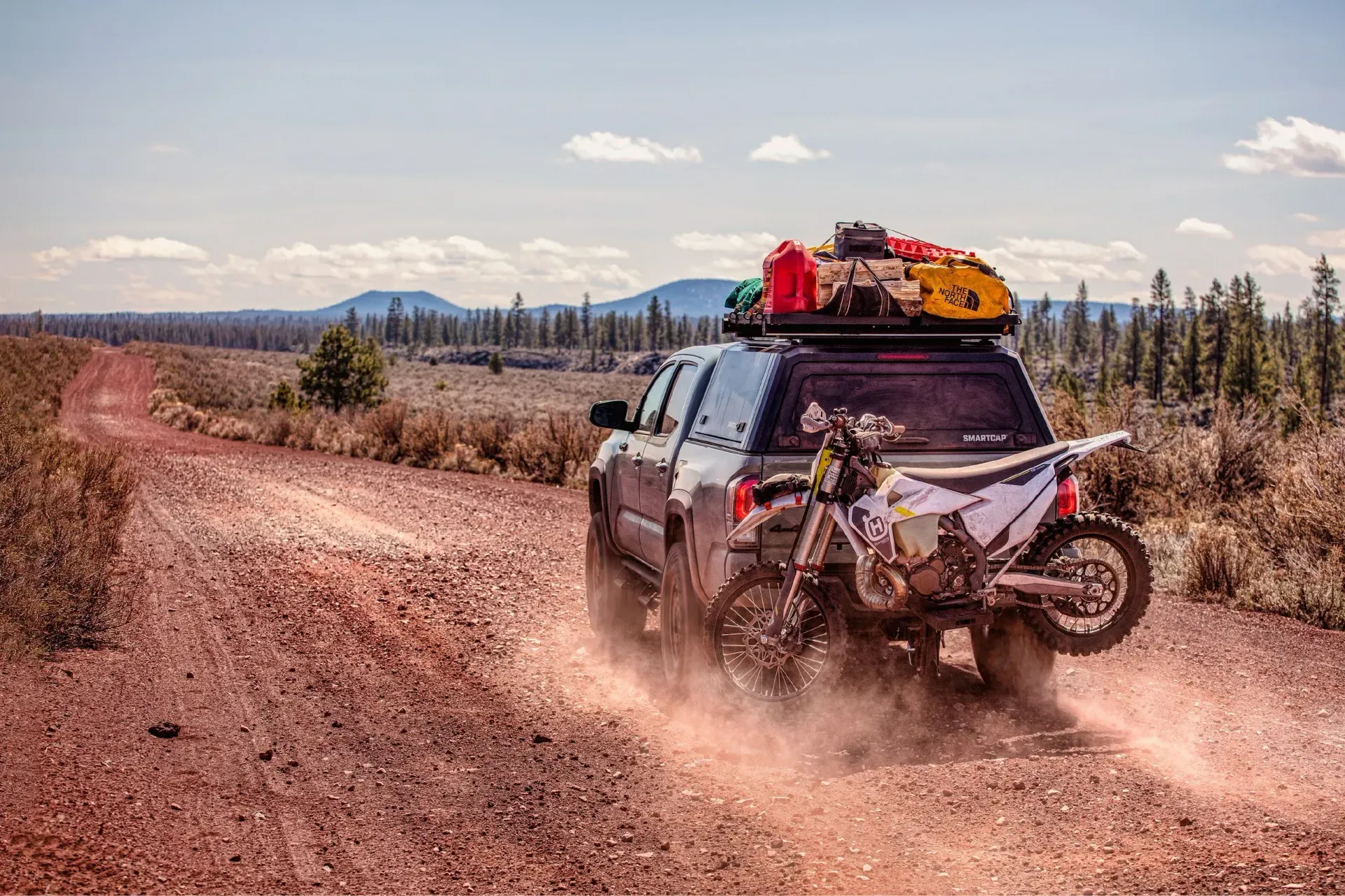 Truck driving on dirt road carrying a motorcycle. Gear on roof. Dust kicking up. Sunny day.