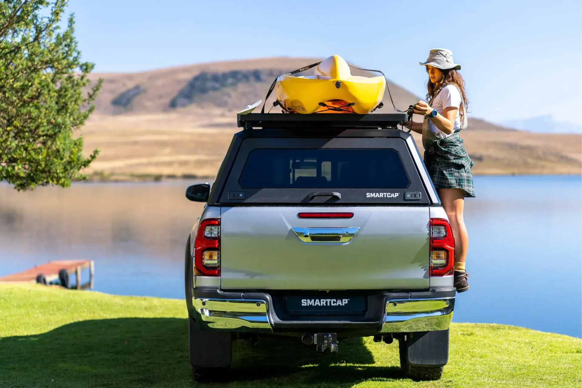 Silver truck with a canoe on top, woman standing at the back, next to a lake.