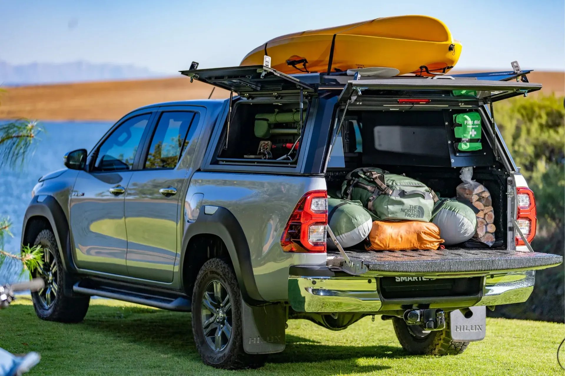 Gray pickup truck with camping gear in the back and a yellow kayak on top, parked on grass near a lake.