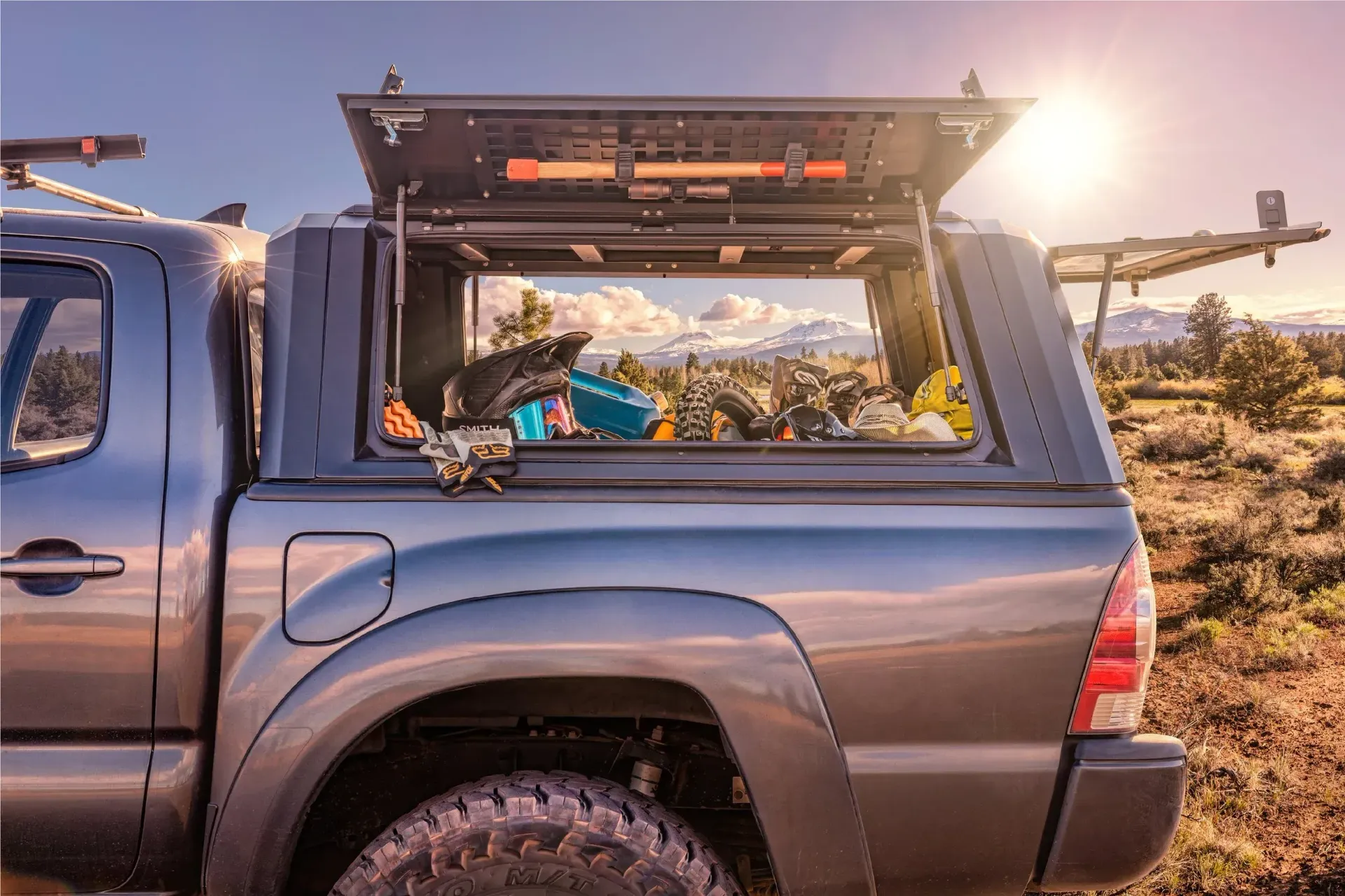Gray pickup truck with an open camper shell filled with camping gear, parked in a sunny outdoor setting with mountains in the background.