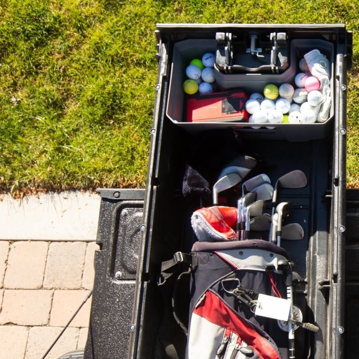 Golf clubs and balls in a truck bed storage compartment, next to green grass and pavement.