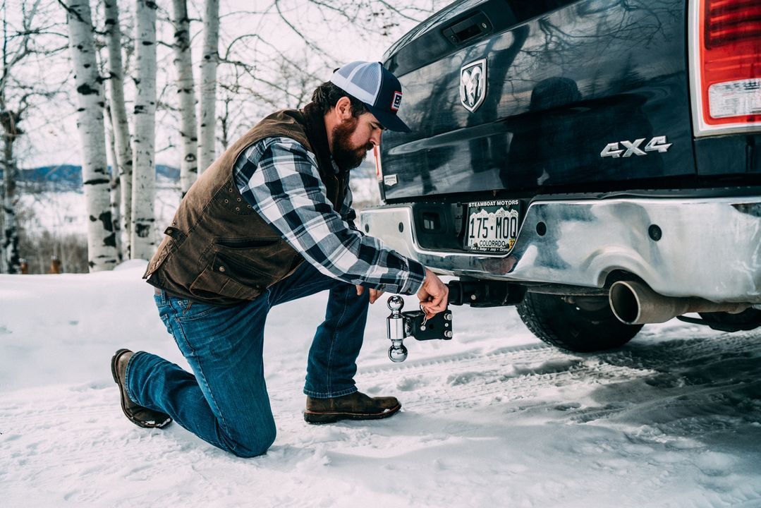 Man kneeling, attaching trailer hitch to truck in snow.
