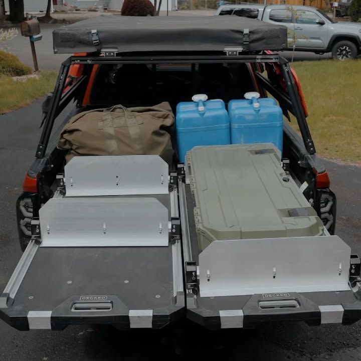 Rear view of a truck bed with slide-out drawers, water containers, and camping gear. Black, gray, and blue.