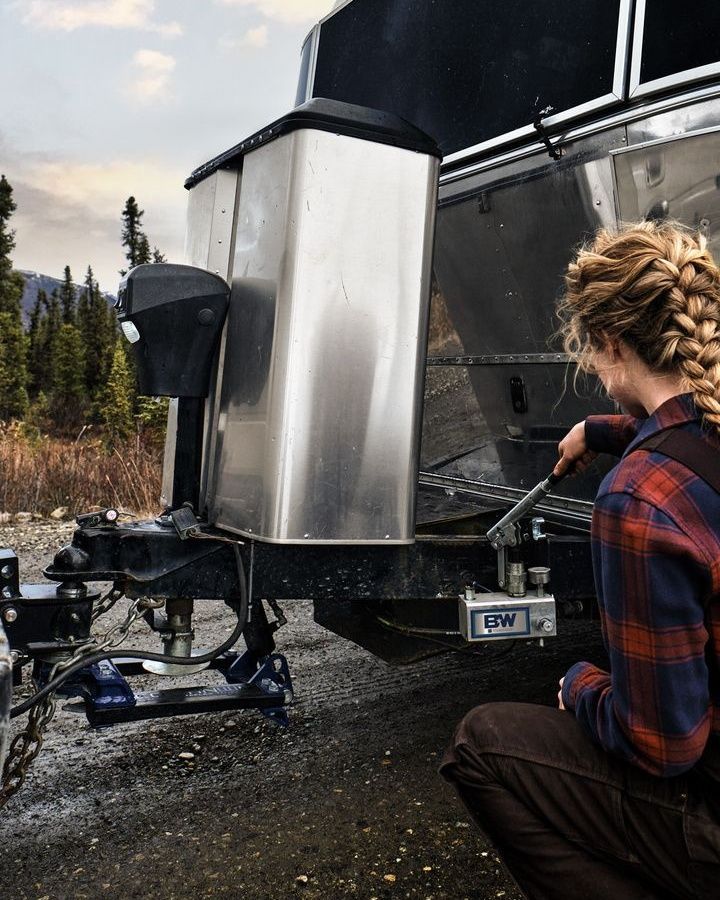 Woman kneeling, adjusting trailer hitch on an Airstream trailer outdoors, with mountains in the background.