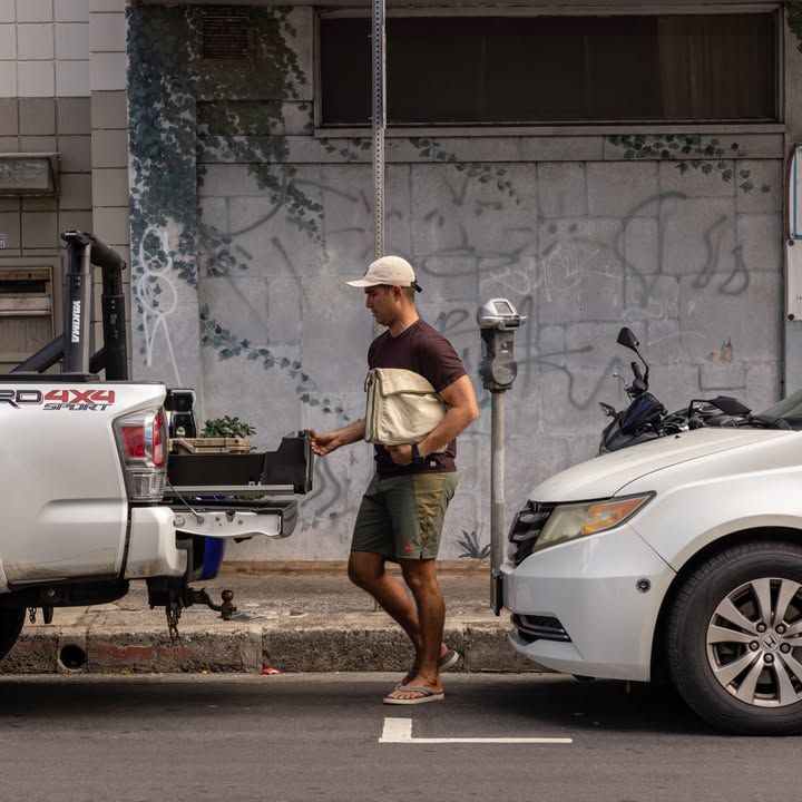 Man unloading items from a truck, standing next to a parked white car on a city street.