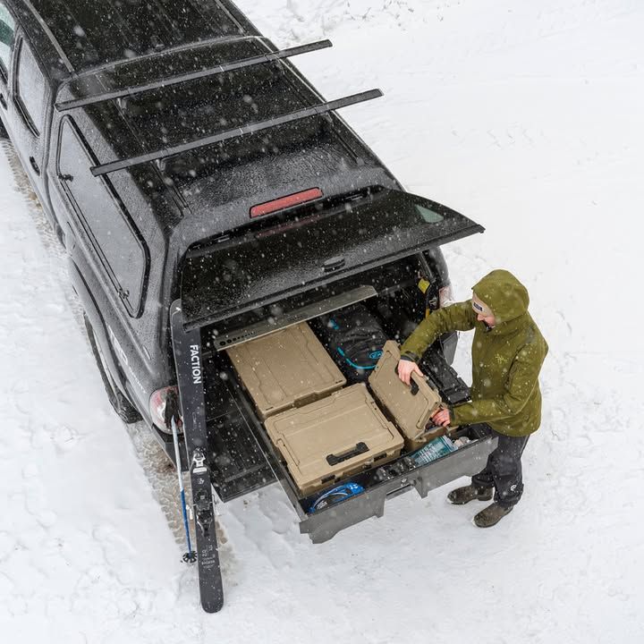 Person loads cargo boxes into truck bed covered by a black canopy in snowy setting.