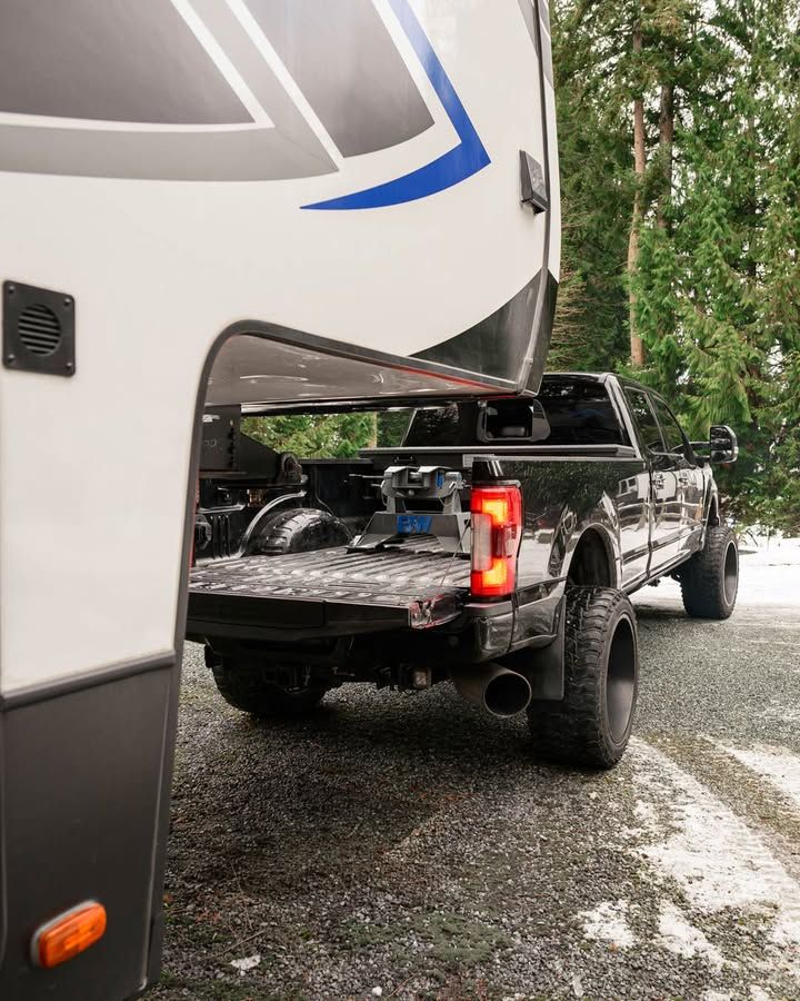 Black truck hitched to a fifth-wheel RV, parked on snowy ground near trees.