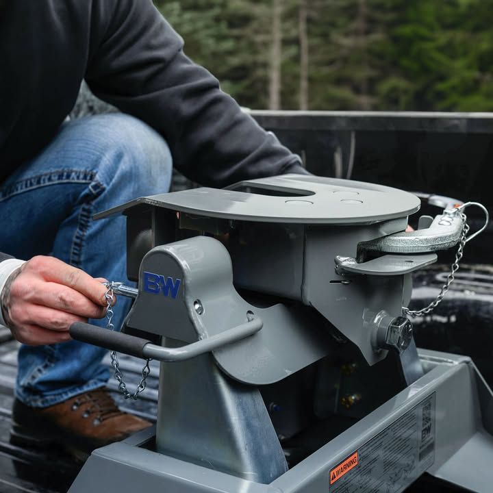 Person attaching a safety chain to a grey fifth-wheel trailer hitch in a truck bed.