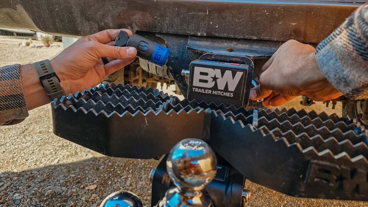 Hands installing a trailer connector near a black hitch and step, outdoors.