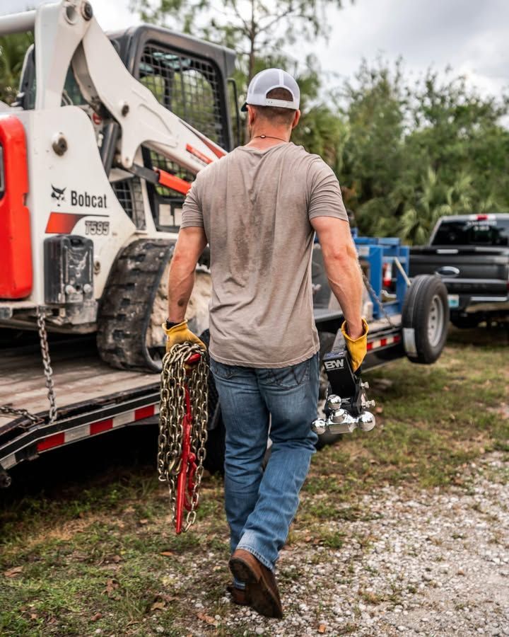 Man carrying chains, walking away from a Bobcat and trailer in a grassy area.
