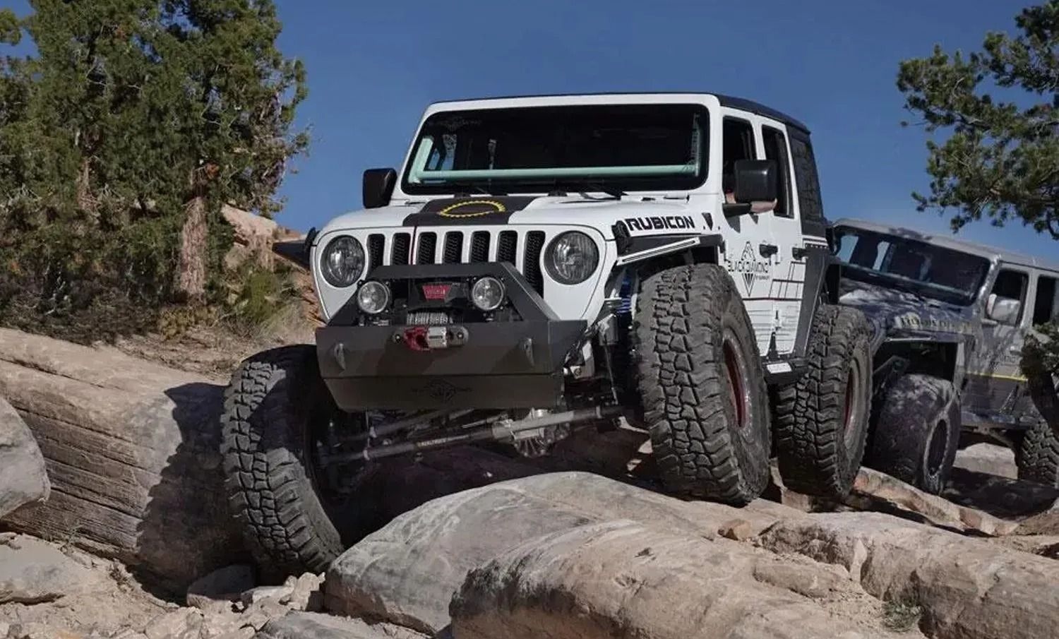 White Jeep truck climbing over large rocks on a sunny, outdoor trail.