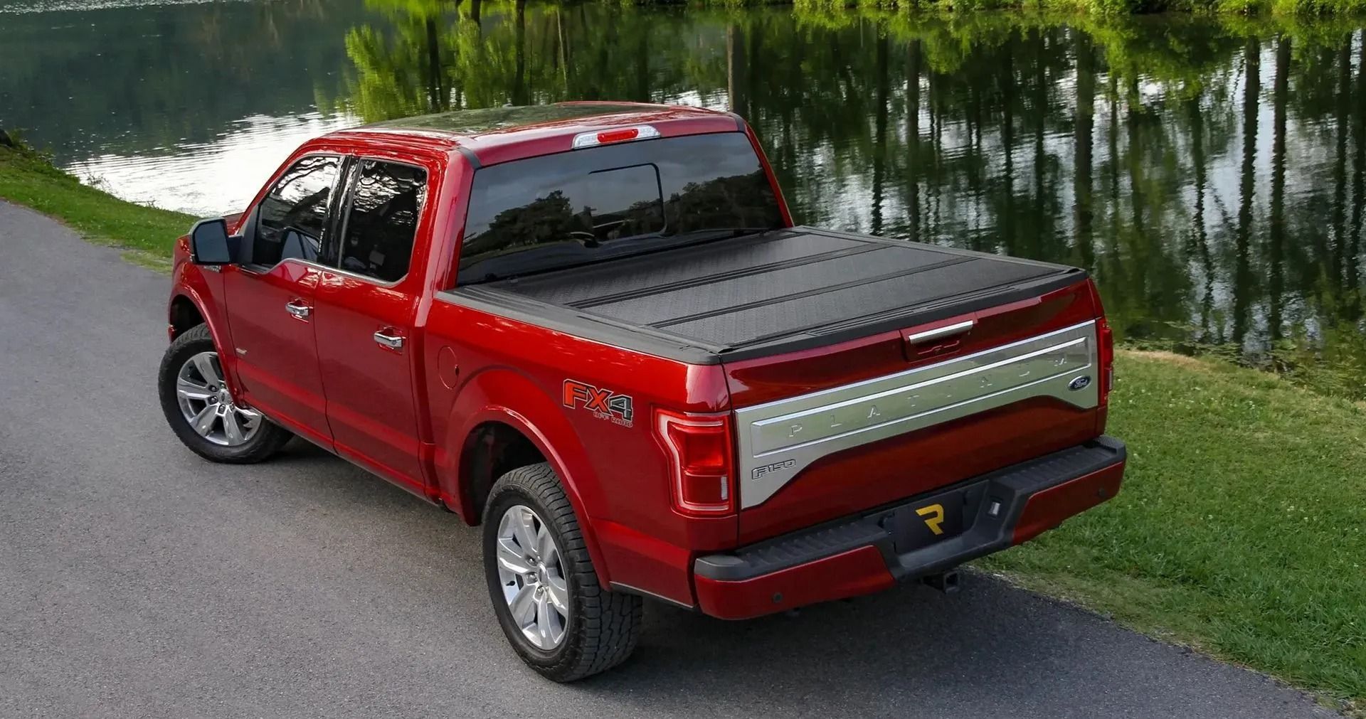 Red Ford F-150 pickup truck with black bed cover parked on a road beside water and grass.