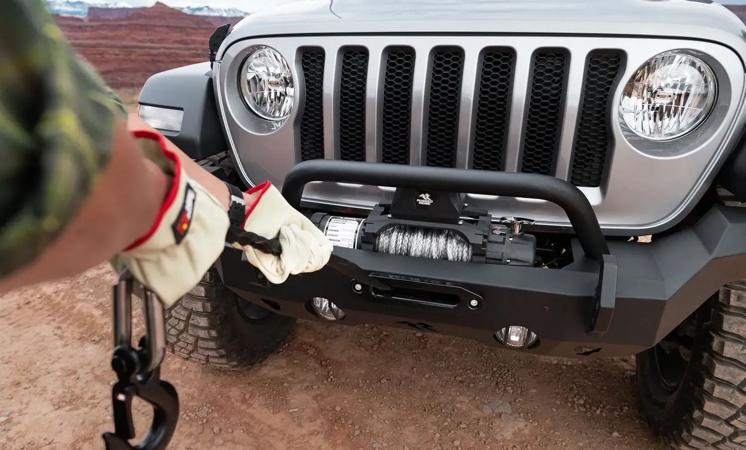 Person in work gloves attaches a hook to a Jeep's winch in a desert setting.