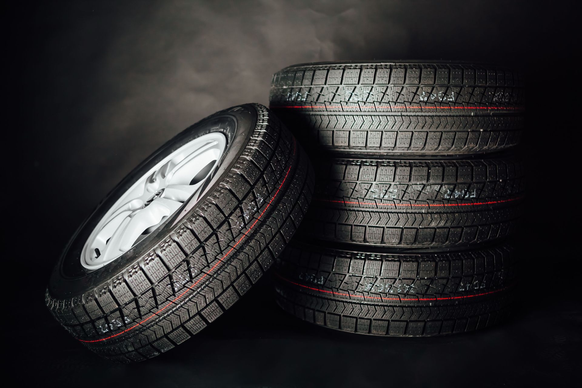 Four black tires stacked, one leaning. Silver wheel, wet appearance, dark background.