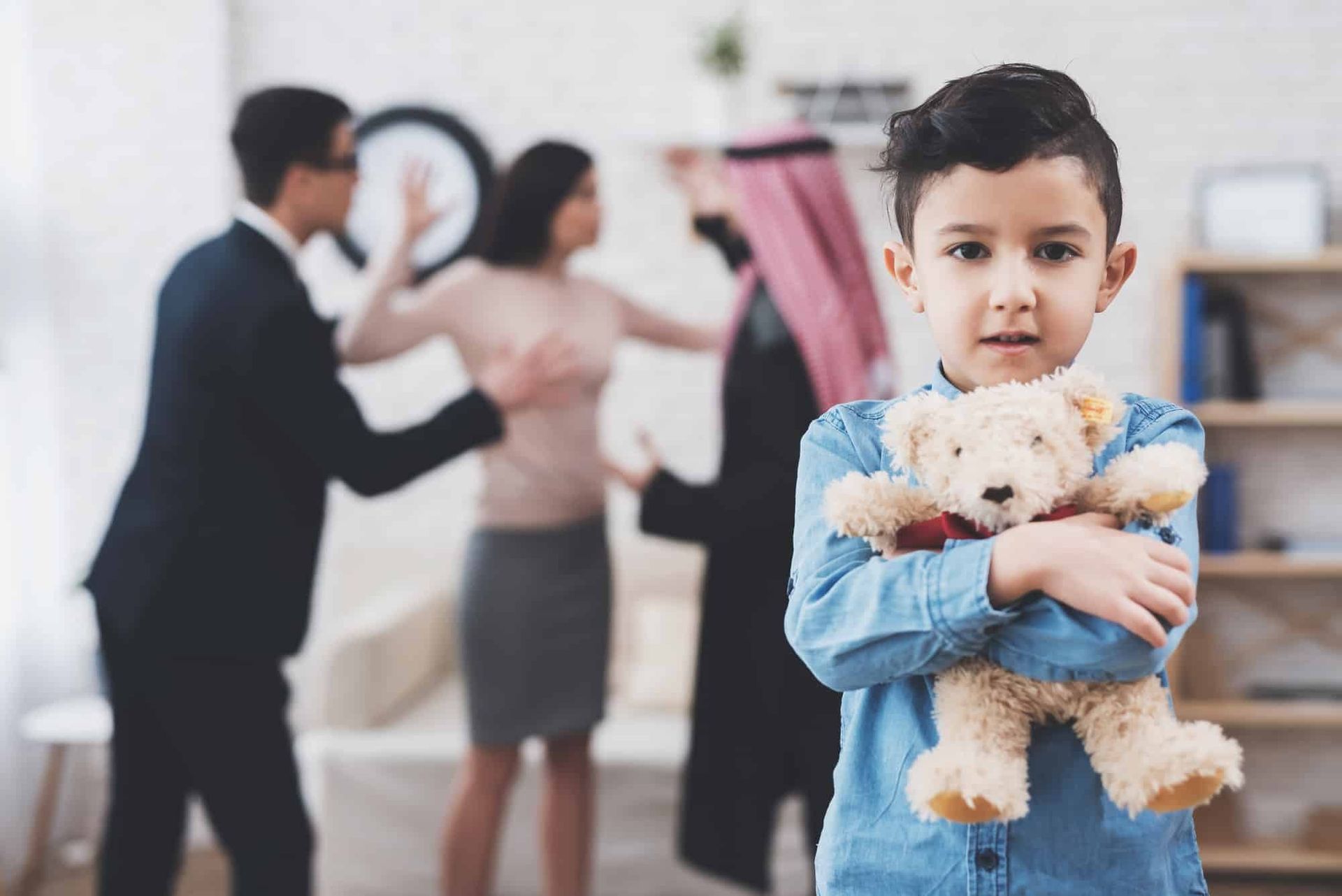 Boy in blue shirt holding a teddy bear, with blurred adults in the background at home.