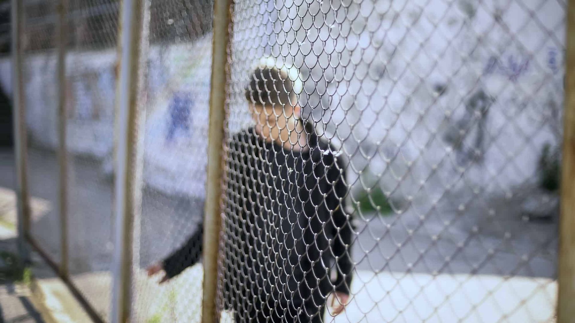 Person standing behind a chain-link fence outdoors, wearing a black jacket.