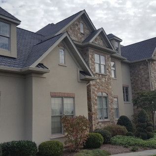 Suburban house exterior with gray stone and tan siding, gabled roof, windows, and manicured shrubs