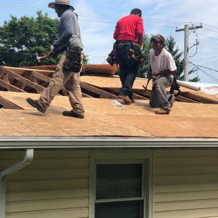 Workers repairing a house roof, with one kneeling and two standing on exposed plywood.