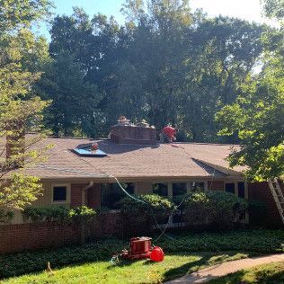 Workers on a house roof under trees, with a lawn mower in the foreground.