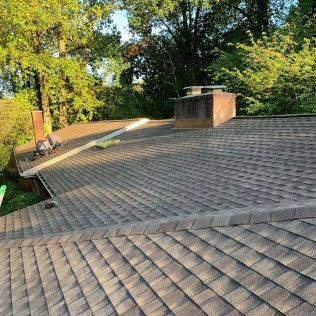 Rooftop with gray shingles, a brick chimney, and trees in the background in warm sunlight