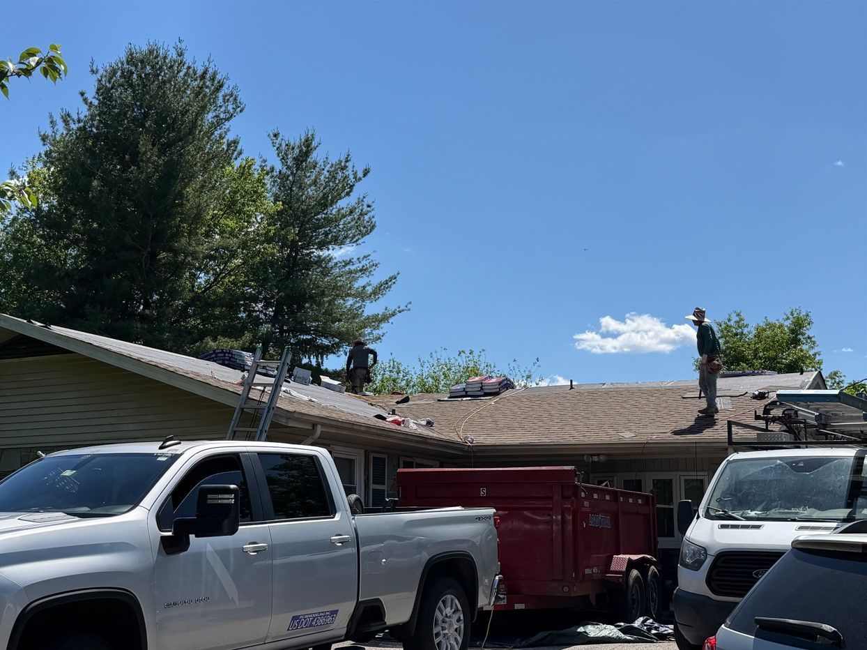 Two pickup trucks and a red trailer parked beside a house with a damaged roof under a clear blue sky