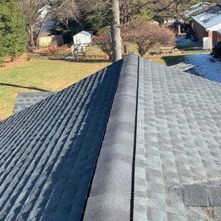 View from a roof ridge with gray asphalt shingles, overlooking a yard and trees below.