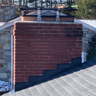 Red brick chimney with black cap on a gray shingled roof against a stone wall.