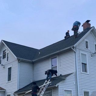 Workers on a white house roof repairing shingles, with a ladder leaning against the side.