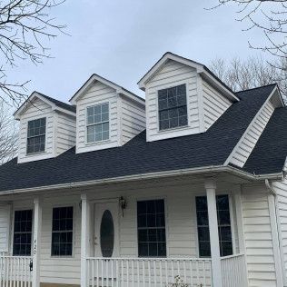 White house with dark roof and three dormer windows, front porch, and bare tree branches overhead