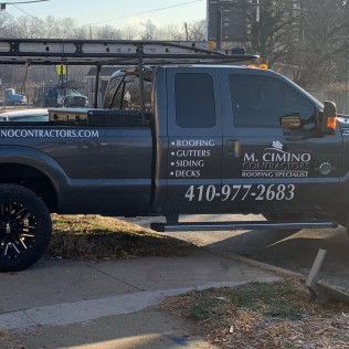 Dark blue contractor pickup truck with ladder rack parked on a sidewalk, company logos and phone number visible