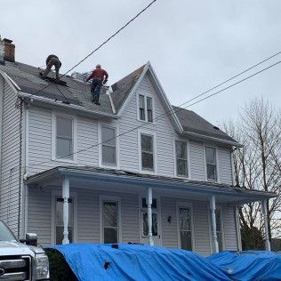 Workers on a gray house roof near the ridge, with a blue tarp covering the porch below.