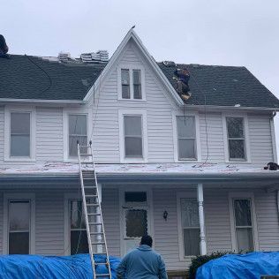 Workers on a roof and ladder repairing a gray house with blue tarp covering part of the porch.