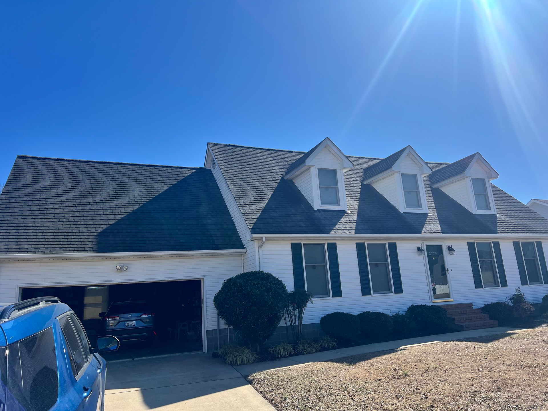 Suburban white house with blue roof, open garage, and a blue car in the driveway under a bright sky