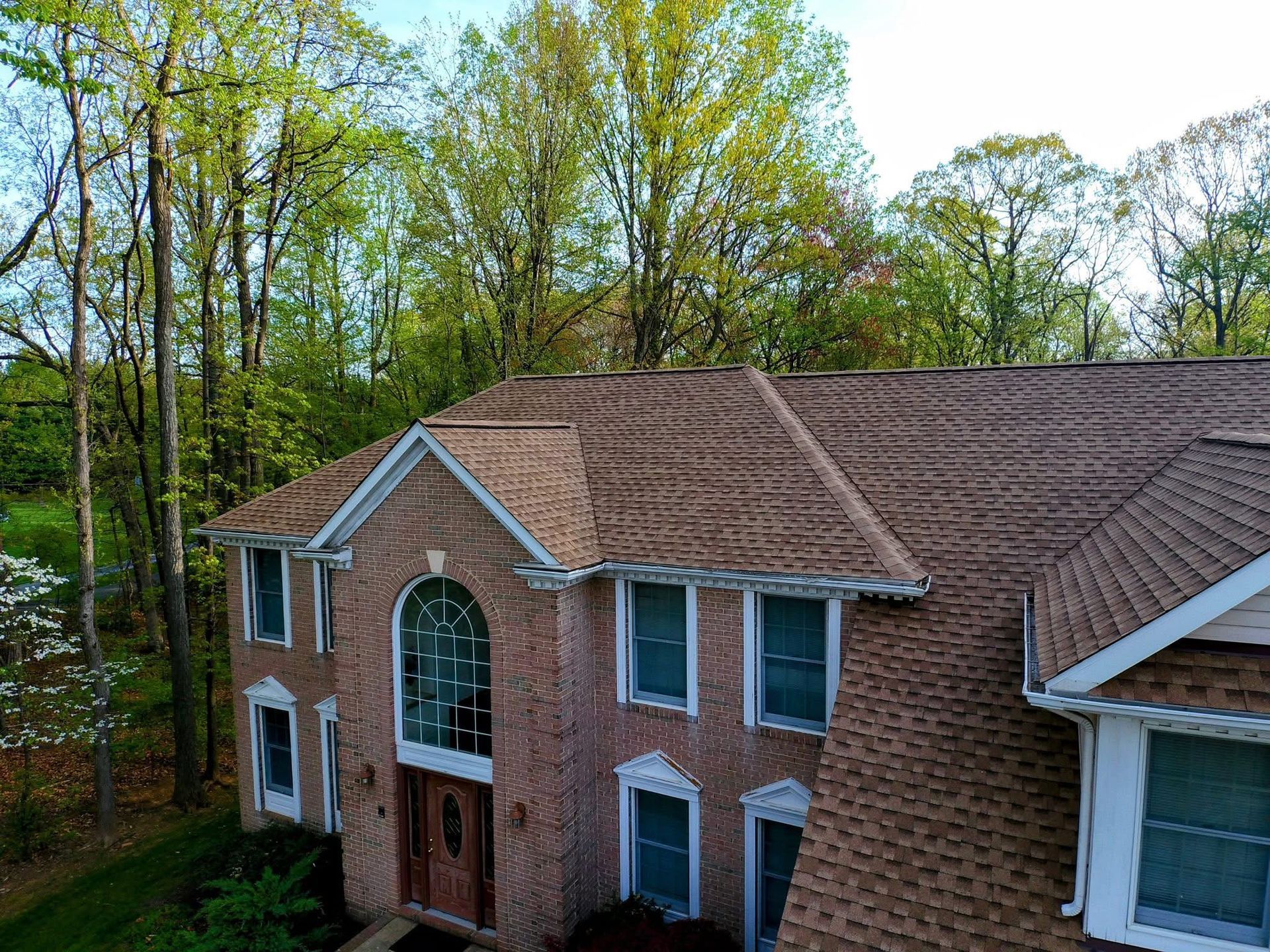 Brick two-story house with brown roof and white-trimmed windows, surrounded by tall trees.