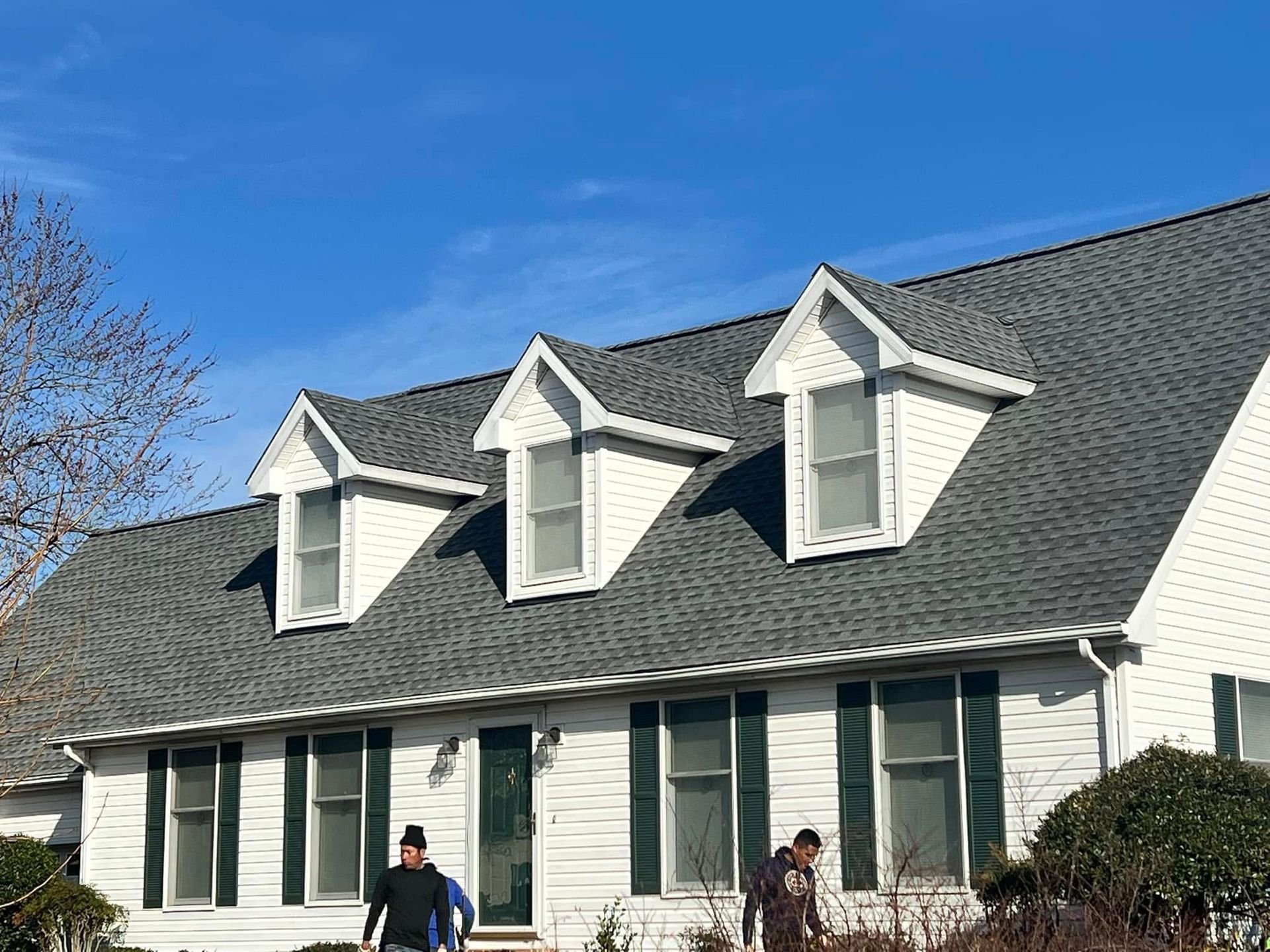 White house with dark roof and dormer windows under a clear blue sky; two people walk in front.