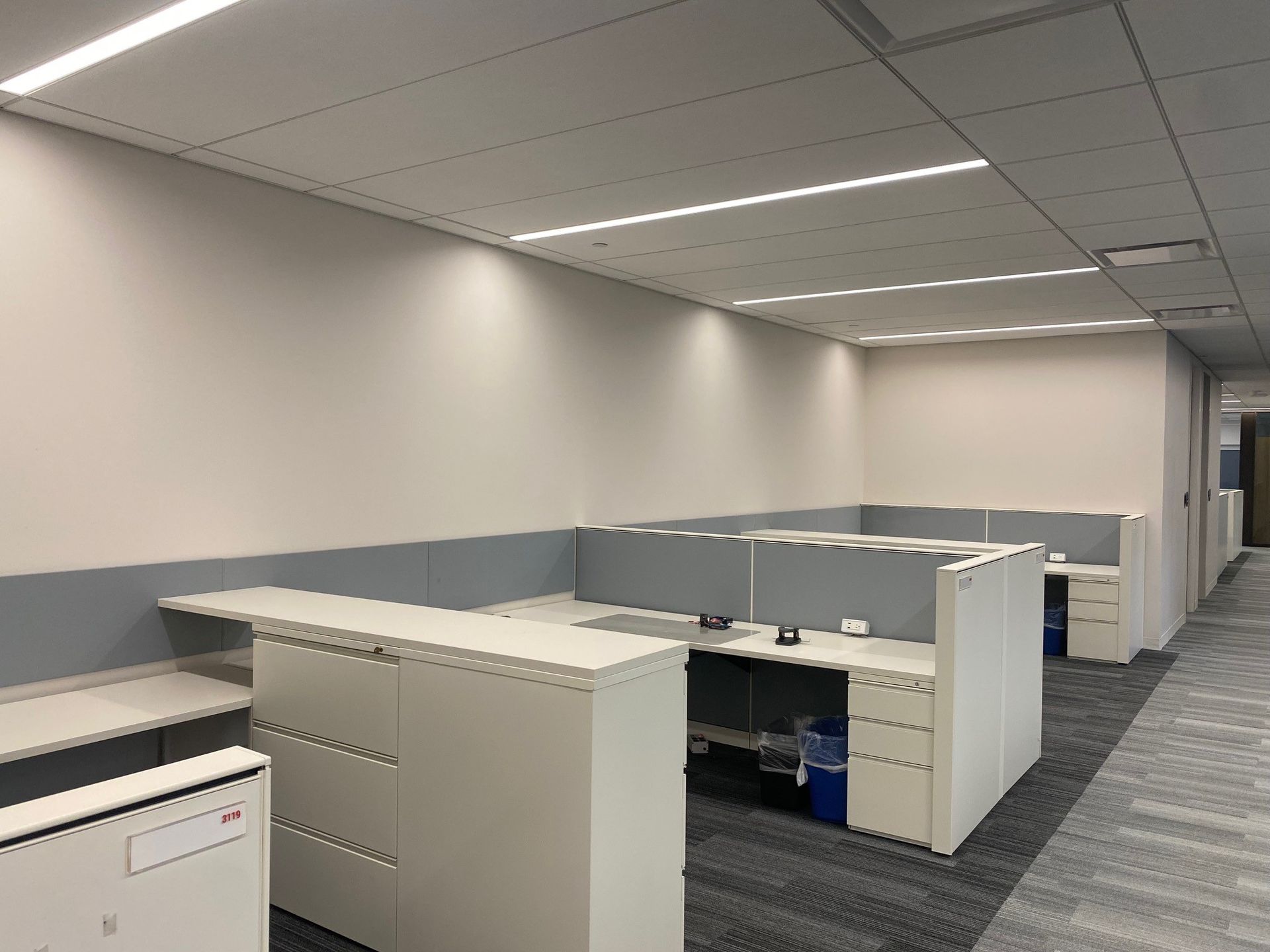 Empty office cubicles in a gray, carpeted workspace with fluorescent ceiling lights
