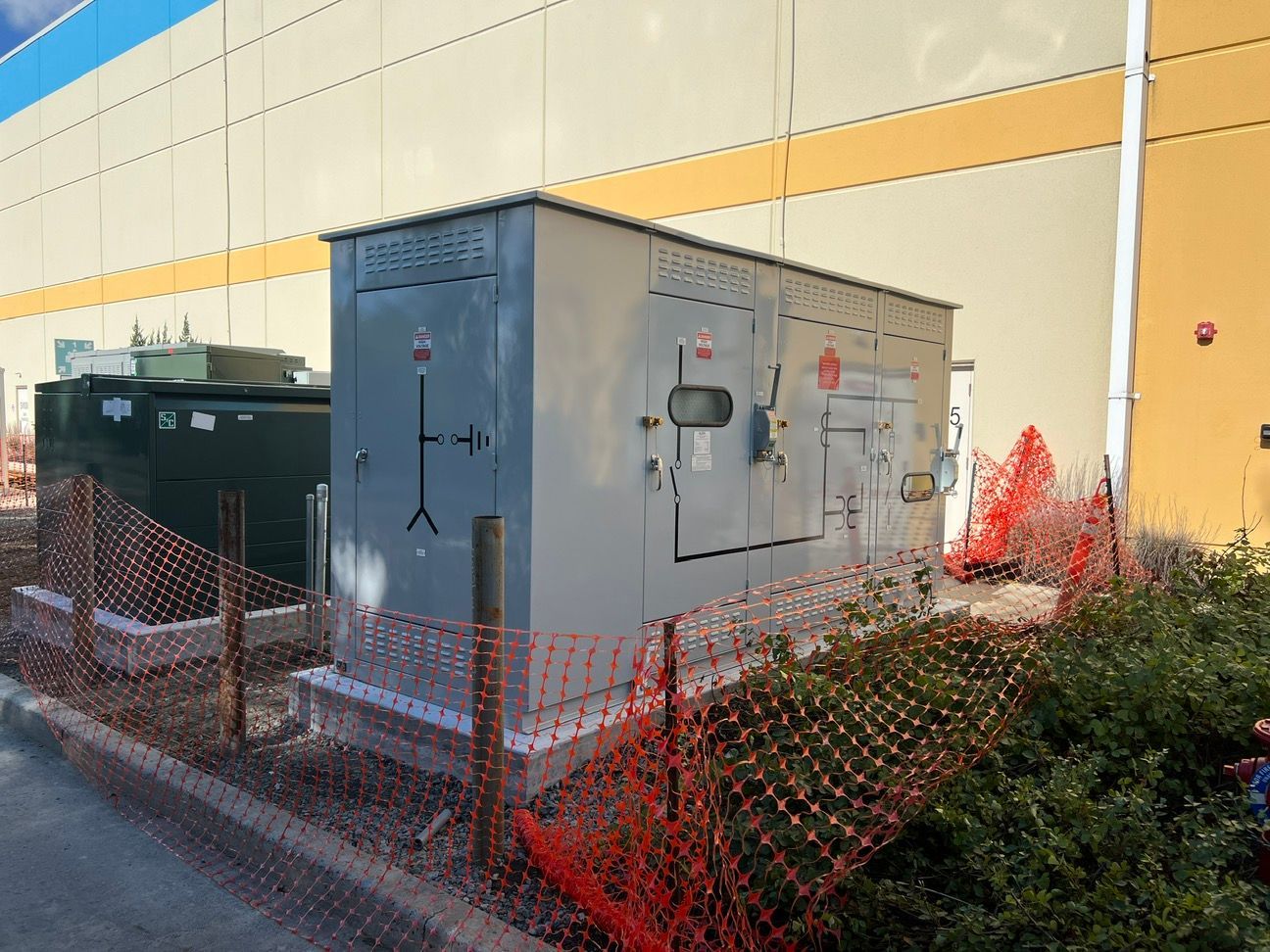 Large gray electrical generator enclosure beside a building, surrounded by orange safety fencing.