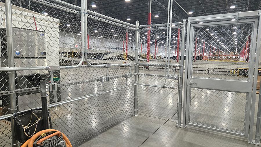 Wire-mesh industrial walkway with metal railings, concrete floor, and overhead lights in a warehouse-like space