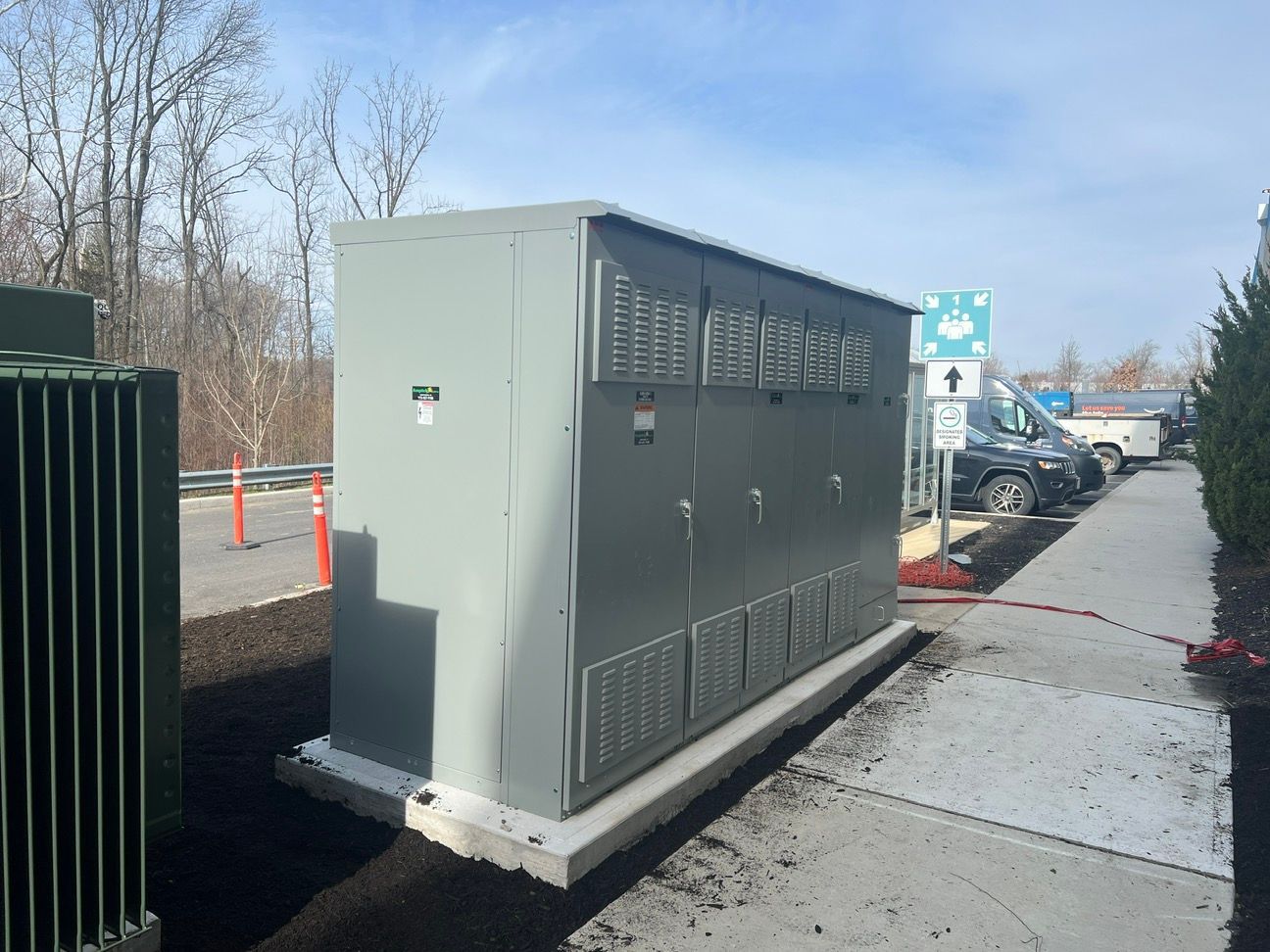 Gray electrical transformer cabinet beside a sidewalk in a parking lot, with cars and trees in the background